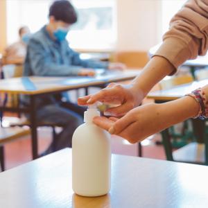 Youth sitting at desk with COVID mask while close up of another using the hand sanitizer